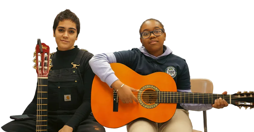 Two young musicians sitting side by side with acoustic guitars, one holding the guitar ready to play and the other resting her guitar upright.